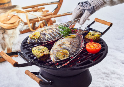 Auf einem Grill im Freien werden zwei Fische in Grillkörben zusammen mit Artischocken, einer halben Tomate und frischen Kräutern, darunter Rosmarin, gegrillt. Im Hintergrund sind ein Schlitten, ein Lederrucksack und ein Fellbezug auf Schnee zu sehen.