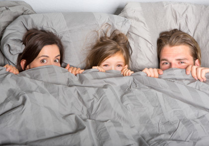 Eine Frau, ein Kind und ein Mann liegen im Bett und schauen überrascht über die Bettdecke, die sie bis zur Nase hochgezogen haben.