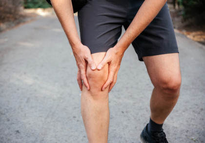 Eine Person in kurzer Hose legt ihre Hände um ihr schmerzendes Knie während eines Spaziergangs auf einem Weg.