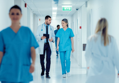 Mehrere Ärzte und Pflegekräfte gehen einen hell erleuchteten Krankenhausflur entlang. Ein Arzt in weißem Kittel und eine Krankenschwester in blauer Uniform führen ein Gespräch, während sie gehen. Es sind weitere medizinische Fachkräfte zu sehen.