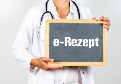 Ein Arzt oder eine Ärztin in weißem Kittel hält eine Tafel mit der Aufschrift "e-Rezept". Ein Stethoskop hängt um den Hals der Person.