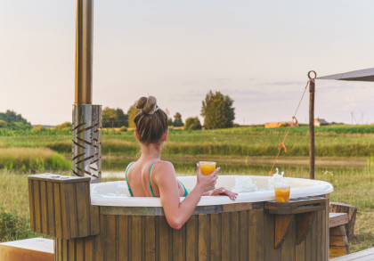 Eine Frau sitzt in einem Whirlpool im Freien mit Blick auf eine grüne Landschaft. Sie trägt ein türkisfarbenes Bikinioberteil und hält ein Glas Orangensaft in der rechten Hand. Ein weiteres Glas steht am Rand des Whirlpools.