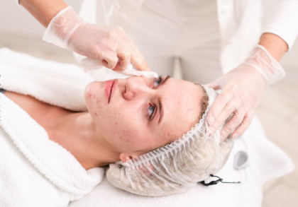 Eine Frau liegt in einem Kosmetikstudio und erhält eine Gesichtsbehandlung. Sie trägt ein weißes Handtuch um die Schultern und ein Haarnetz, während die Kosmetikerin mit Handschuhen sanft ihr Gesicht reinigt.