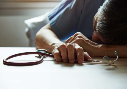Ein erschöpfter Arzt liegt mit einem Stethoskop in der Hand auf einem Tisch. Der Arzt trägt eine blaue Krankenhausschürze und scheint nach einem langen Arbeitstag eingeschlafen zu sein. Die Szene vermittelt Müdigkeit und Überarbeitung.