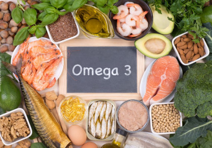 Eine Tafel mit der Aufschrift "Omega 3" steht im Zentrum, umgeben von Lebensmitteln, die reich an Omega-3-Fettsäuren sind, darunter Avocado, Lachs, Krabben, Nüsse, Olivenöl, Fisch, Eier, Walnüsse und grünem Gemüse wie Brokkoli.