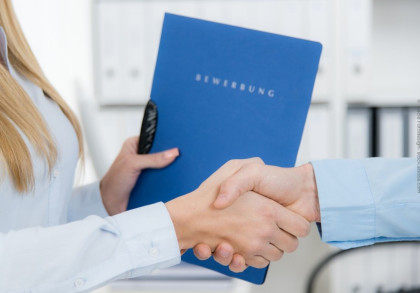 Person in weißem Hemd, die einer anderen Person in blauem Hemd die Hand schüttelt. Die Person im weißen Hemd hält eine blaue Bewerbungsmappe mit dem Wort "BEWERBUNG" darauf. Im Hintergrund sind unscharfe, weiße Büromöbel zu sehen.