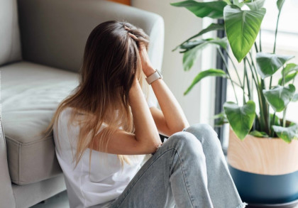 Eine Frau sitzt auf dem Boden, lehnt sich gegen ein Sofa und hält ihren Kopf in ihren Händen. Neben ihr steht eine grüne Zimmerpflanze. Die Szene wirkt nachdenklich und melancholisch.