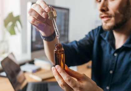 Ein Mann hält eine braune Glasflasche und verwendet eine Pipette, um Flüssigkeit aus der Flasche zu entnehmen. Er arbeitet an einem Schreibtisch mit einem Laptop und anderen Büroartikeln im Hintergrund.