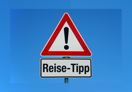 Ein dreieckiges Verkehrsschild mit rotem Rand und schwarzem Ausrufezeichen auf weißem Hintergrund. Darunter ein rechteckiges Schild mit der Aufschrift "Reise-Tipp" in schwarzer Schrift. Der Hintergrund ist ein klarer blauer Himmel.