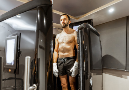 Ein Mann in Shorts und Handschuhen steht in einer Kryokammer in einem modernen Wellness-Zentrum. Er bereitet sich auf eine Kältetherapie vor, umgeben von futuristischer Ausrüstung und gedämpfter Beleuchtung.