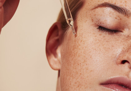 Nahaufnahme eines Gesichts mit Sommersprossen, während eine Pipette ein Hautpflege-Serum auf die Wange aufträgt. Die Person hat geschlossene Augen, was Entspannung und Pflege symbolisiert.