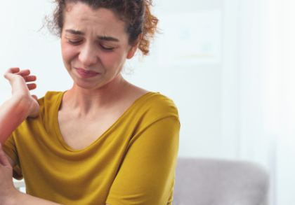 Frau in gelbem Oberteil kratzt sich am geröteten Unterarm, sichtbar von Hautreizungen betroffen. Das Bild zeigt typische Symptome von Neurodermitis, wie Rötungen und Juckreiz. Im Hintergrund ein heller Wohnraum mit Pflanzen und Pflegeprodukten.