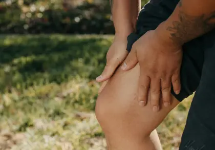 Eine Person umfasst ihr schmerzendes Knie mit beiden Händen. Es wird der Eindruck von Gelenk- und Muskelschmerzen, nach einer körperlichen Aktivität vermittelt. Der Hintergrund ist unscharf und zeigt eine Wiese im Sonnenlicht.