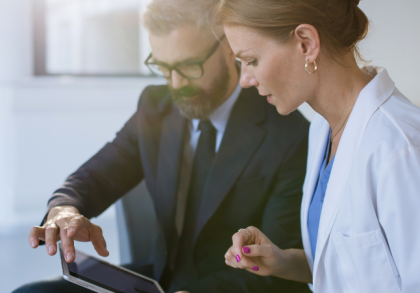 Ärztin im weißen Kittel bespricht mit einem Anzugträger am Tablet digitale Marketingstrategien zur Patientenakquise in einer modernen Praxisumgebung.
