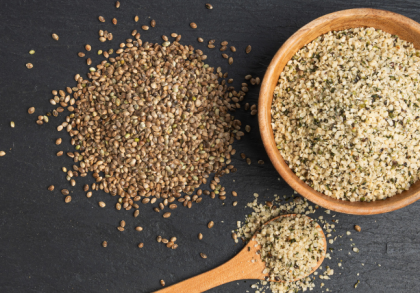 Ungeschälte Hanfsamen und geschälte Hanfkerne in einer Holzschale sowie auf einem Löffel, arrangiert auf dunkler Steinoberfläche – Darstellung verschiedener Formen von Hanfsamen für Ernährung und Lebensmittelverarbeitung.