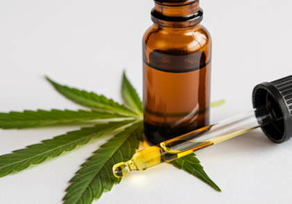 Braune CBD-Ölflasche mit Pipette und goldfarbenem Öl auf grünen Hanfblättern vor hellem Hintergrund.
