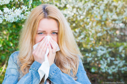 Eine blonde Frau mit langer Haare niest in ein Taschentuch. Sie trägt eine Jeansjacke und steht vor einem blühenden Baum im Freien.