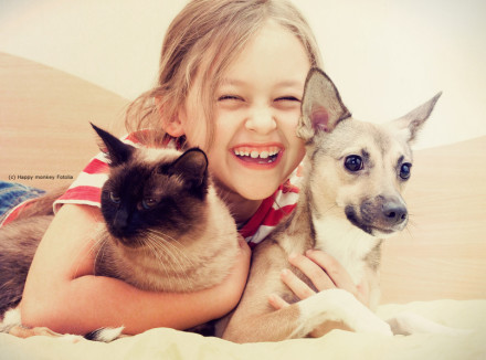 Ein lachendes Kind hält eine braune Katze und einen kleinen Hund in den Armen. Das Kind trägt ein rot-weiß gestreiftes T-Shirt. Die Szene wirkt fröhlich und warm.