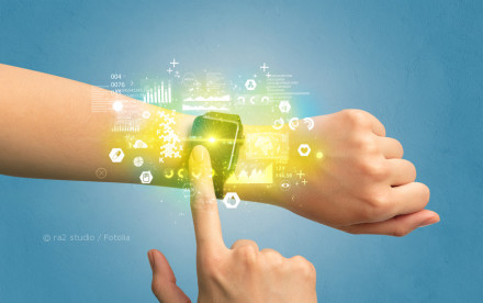 Eine Hand zeigt auf eine Smartwatch, die an einem Handgelenk getragen wird. Von der Smartwatch strahlen holografische Symbole und Datenanzeigen in leuchtenden Farben. Der Hintergrund ist blau.