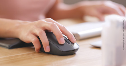 Eine Hand bewegt eine Computermaus auf einem Holztisch. Im Hintergrund sind unscharf eine Tastatur und andere Objekte zu sehen.