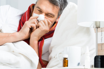 Ein Mann liegt krank im Bett, trägt einen roten Schal und hält ein Taschentuch an seine Nase. Auf dem Nachttisch neben ihm stehen eine Tasse, eine kleine braune Flasche und ein Nasenspray.