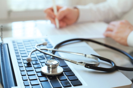 Ein Stethoskop liegt auf der Tastatur eines Laptops, während im Hintergrund eine Hand mit einem Stift auf einem Blatt Papier schreibt. Medizinische oder gesundheitliche Atmosphäre, möglicherweise in einer Arztpraxis oder einem Büro.