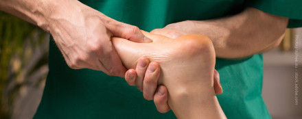 Nahaufnahme von einer Person in grüner Kleidung, die eine Fußmassage durchführt, indem sie den Fuß einer anderen Person mit beiden Händen hält.