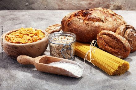 Eine Auswahl an kohlenhydratreichen Lebensmitteln in einem rustikalen Arrangement. Zu sehen sind ein Laib Brot, zwei Brötchen, ein Haufen unverkochter Spaghetti, eine Schale mit Cornflakes, ein Glas mit Haferflocken und eine hölzerne Schaufel mit Mehl.