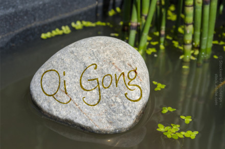 Ein großer, glatter Stein liegt im Wasser, auf dessen Oberfläche das Wort "Qi Gong" geschrieben ist. Im Hintergrund sind Bambusrohre und schwimmende Pflanzenblätter zu sehen. Die Umgebung vermittelt eine ruhige und friedliche Atmosphäre.