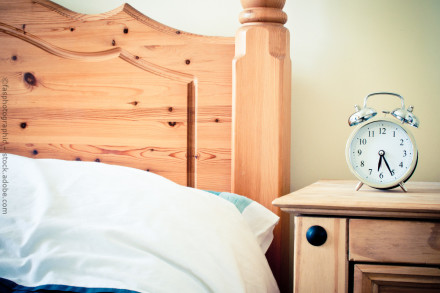 Holzbett mit Kopfteil und weißer Bettwäsche daneben ein hölzerner Nachttisch mit einem silbernen Wecker.
