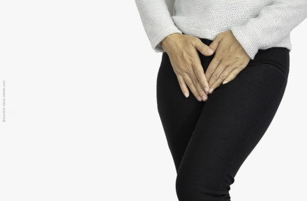 Eine Person in einem weißen Pullover und schwarzen Hosen hält beide Hände in der Nähe des Unterbauchs.