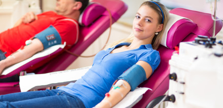 Eine junge Frau sitzt auf einem medizinischen Liegestuhl und spendet Blut. Im Hintergrund ein weiterer Blutspender. Beide haben Blutdruckmanschetten am Arm und sind mit medizinischen Geräten verbunden.