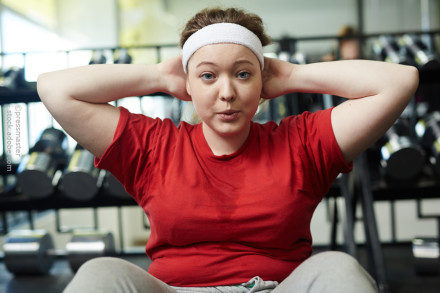 Eine Frau in einem roten T-Shirt und mit einem weißen Stirnband macht Bauchmuskelübungen im Fitnessstudio. Im Hintergrund sind Hanteln zu sehen.