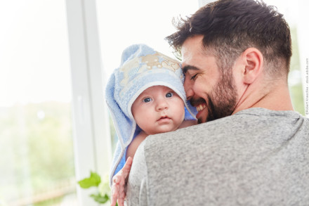 Ein Mann mit Bart hält ein Baby auf seinem Arm. Das Baby hat ein blaues Handtuch mit Kapuze auf dem Kopf und schaut neugierig in die Kamera, während der Mann lächelnd das Baby betrachtet. Sie befinden sich in einem hellen Raum vor einem Fenster.