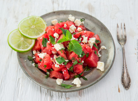 Ein Teller mit Wassermelonen-Feta-Salat, garniert mit frischer Minze und zwei Limettenscheiben auf der Seite. Rechts vom Teller liegt eine antike Gabel auf einem weißen Holztisch.