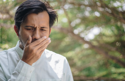 Ein Mann im Freien hält sich die Hand vor den Mund und hat einen besorgten Gesichtsausdruck. Er trägt ein helles Hemd, und im Hintergrund sind verschwommene grüne Bäume zu sehen.