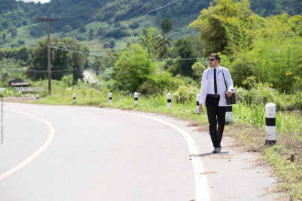 Ein Mann im Arztkittel mit einer Tasche und einer Sonnenbrille geht am Straßenrand einer ländlichen Straße entlang, umgeben von grüner Landschaft. Im Hintergrund sind Hügel und Bäume zu sehen. Es ist ein sonniger Tag mit klarem Himmel.