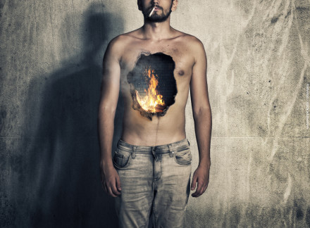 Ein Mann steht mit nacktem Oberkörper vor einer Betonwand, während er eine Zigarette raucht. Ein großes Loch mit brennendem Feuer ist in der Mitte seiner Brust dargestellt, wodurch es so aussieht, als ob sich das Feuer in seinem Inneren befindet.
