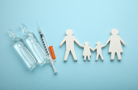 Zwei Ampullen mit Flüssigkeit und eine Spritze liegen auf blauem Hintergrund. Neben den Medikamenten sind Holzfiguren, die eine Familie darstellen, abgebildet. Die Familie besteht aus zwei Erwachsenen und zwei Kindern, die sich an den Händen halten.
