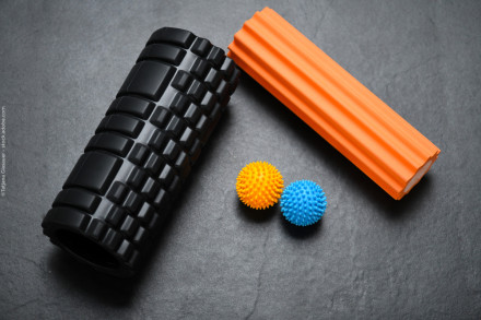 Bildbeschreibung in Deutsch: Zwei Massagerollen, eine schwarze und eine orangefarbene, liegen auf einem dunklen Hintergrund. Neben ihnen liegen zwei stachelige Massagebälle, einer gelb und einer blau.