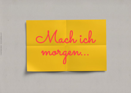 Das Bild zeigt ein gelbes Blatt Papier mit dem handgeschriebenen Text "Mach ich morgen..." in pinker Farbe. Das Blatt ist leicht gefaltet und vor einem grauen Hintergrund platziert.