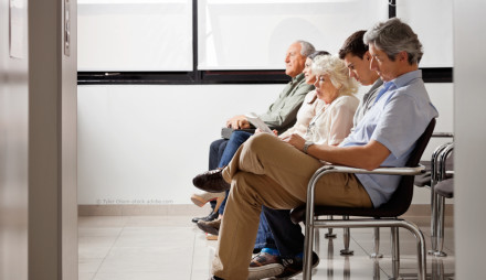 Eine Gruppe von vier Personen sitzt auf Stühlen in einem Wartezimmer. Sie sind in einer Reihe nebeneinander angeordnet und scheinen zu warten. Zwei der Personen lesen, während die anderen zwei geradeaus schauen.