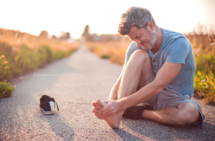 Ein Mann sitzt auf einem ländlichen Weg und hält seinen Fuß mit einem schmerzverzerrten Gesichtsausdruck. Sein linker Schuh liegt neben ihm auf dem Boden. Es ist ein sonniger Tag, und der Weg ist von Gras und Pflanzen gesäumt.