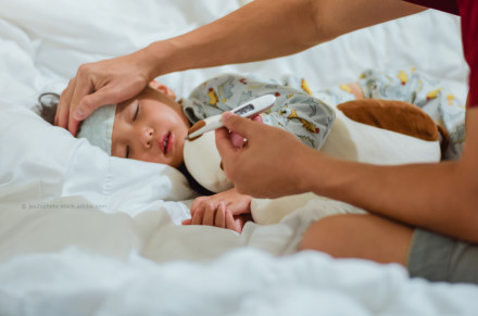 Eine Hand hält ein Thermometer, während die andere Hand die Stirn eines schlafenden Kindes fühlt, das im Bett liegt und ein Kühlkissen auf der Stirn hat. Das Kind trägt einen bunten Schlafanzug mit Tiermotiven.