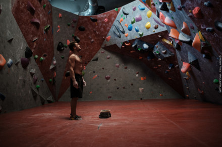 Ein muskulöser Mann steht in einer Kletterhalle mit rot-grau gestreiften Kletterwänden und farbigen Griffen. Er trägt schwarze Shorts und steht auf einem roten Bodenbelag, während ein Kreidebeutel zwischen seinen Füßen liegt.