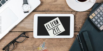 Auf einem Holztisch liegen ein Laptop, eine Armbanduhr, eine Tasse Kaffee, ein Taschenrechner, eine Brille, bunte Büroklammern, ein Notizbuch mit Stift und ein Tablet mit einem Klebezettel darauf. Auf dem Zettel steht in Großbuchstaben "ELTERNZEIT".