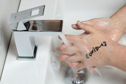 Eine Person wäscht sich die Hände unter fließendem Wasser mit Seife, während das Wort 'COVID-19' auf der Haut geschrieben ist. Der Wasserhahn und das Waschbecken sind ebenfalls sichtbar.