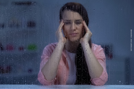 Eine Frau mit langen dunklen Haaren sitzt hinter einem regennassen Fenster. Sie trägt ein rosa Oberteil und stützt ihr Gesicht mit beiden Händen, während sie nachdenklich oder gestresst aussieht.