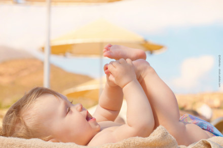 Ein lächelndes Baby, das auf einem Handtuch liegt und mit seinen Füßen spielt. Im Hintergrund sind Sonnenschirme und ein heller, wolkenloser Himmel zu sehen.