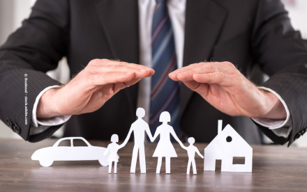 Ein Mann in Anzug und Krawatte hält seine Hände schützend über Papiersilhouetten einer Familie mit zwei Kindern, einem Haus und einem Auto auf einem Tisch.